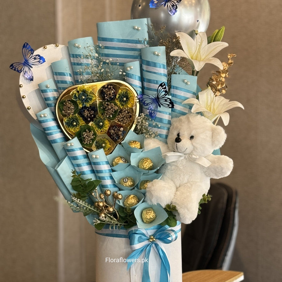 A candy bouquet arrangement featuring an assortment of chocolates including Snickers, Mars, KitKat, Dairy Milk, and Ferrero Rocher, adorned with blue ribbons, artificial blue flowers, and a small white teddy bear.