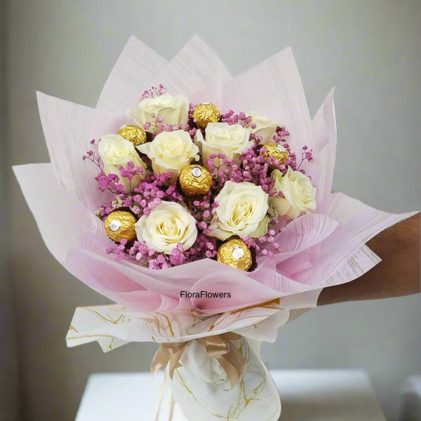 A bouquet of white roses mixed with baby's breath and Ferrero Rocher candies, wrapped in pink and white paper.