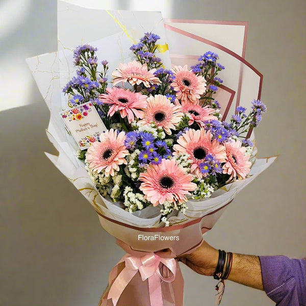 A bouquet of flowers including pink and purple gerberas, baby's breath, and other complementary foliage, wrapped in white paper with a pink ribbon.